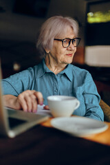 Happy senior woman with glasses sits at a table in front of a laptop Retired woman chatting unaltered