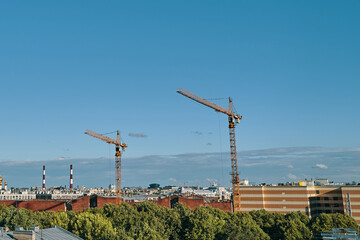 City landscape with copy space. Construction cranes, multi-storey buildings and green trees. Urban area on a sunny summer day. Blue sky with clouds.