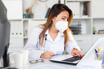 Woman doctor in medical mask is working at laptop in clinic