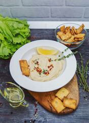 on a gray table on a cutting board, a white plate with hummus, a slice of lemon, spicy seasoning and crackers next to lettuce, sprigs of greens, two bowls, one with olive oil, the other with crackers.