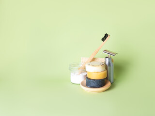 The bamboo toothbrush in glass with toothpowder and steel multiple-use razor in bottle, three natural round soaps and natural wash gourd against the green background. Zero waste bathing concept.