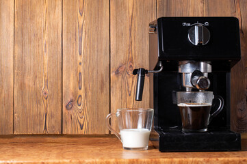 A cup of black coffee and a cup of milk stand near a coffee maker with a manual cappuccino maker. Wood background. Copy space for text, natural