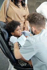 Young dentist in uniform, gloves and protective mask putting napkin on chest of little patient before examination of oral cavity