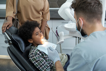 Youthful African boy with open mouth looking at male dentist with instruments before oral check-up