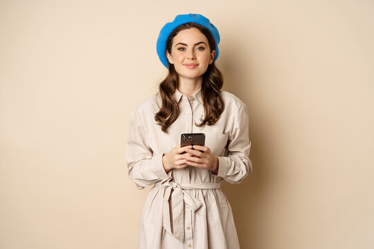 Image of young stylish french woman in trench coat, standing with smartphone, using mobile phone app and smiling, posing against beige background