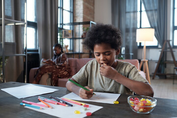 Cute boy eating candies and drawing picture with crayons while sitting by table in living-room against his mother - obrazy, fototapety, plakaty