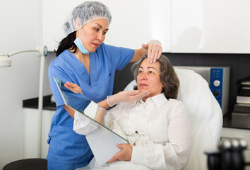 Qualified asian female cosmetologist working with elderly woman client in aesthetic medicine clinic examining face skin before procedure .
