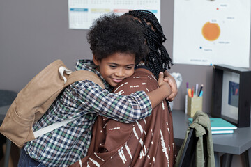 Happy affectionate schoolboy with backpack giving hug to his mother while getting ready for school...