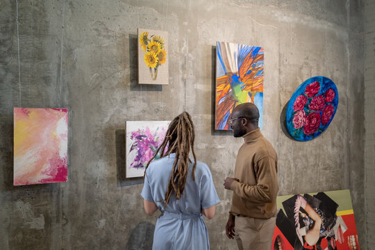 Young intercultural man and woman discussing one of paintings on wall while standing in contemporary art gallery - Powered by Adobe