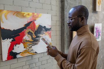 Serious African man with art guide leaflet standing in front of white brick wall with abstract...