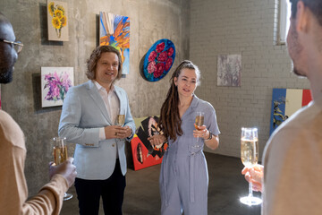 Group of intercultural guests of exhibition with flutes of champagne discussing features of...