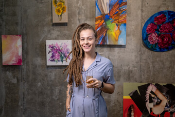 Happy young female with dreadlocks toasting with flute of champagne while standing in front of camera in art gallery