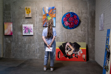 Back view of young woman standing in front of wall with artwork collection and looking at one of...