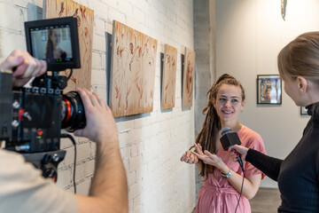 Happy young female guide of art gallery giving interview to journalists during presentation of new artwork collection - obrazy, fototapety, plakaty