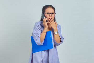 Portrait of cheerful young Asian woman laughing while talking on mobile phone with friends about something funny while holding document folder isolated on purple background