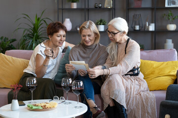 Mature women watching old photos together on the sofa, they drinking wine and remembering past moments during meeting at home