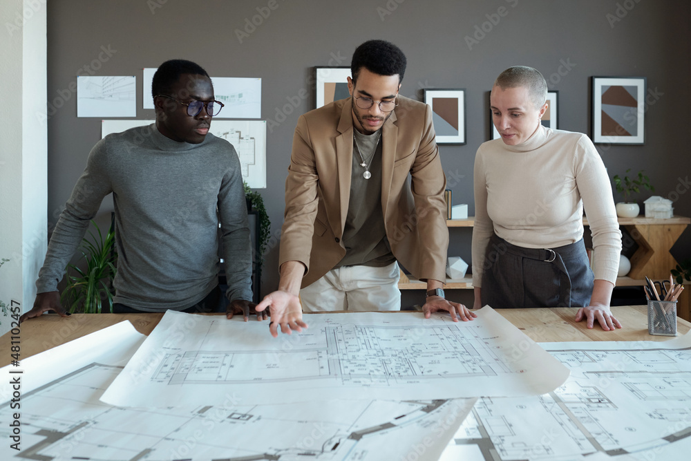 Poster One of three coworkers making presentation of large sketch on blueprint and explaining it to intercultural colleagues