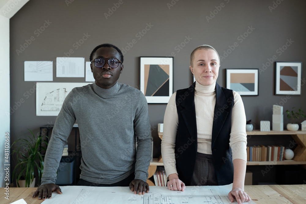 Poster two confident intercultural engineers in casualwear standing by workplace and discussing sketch on b