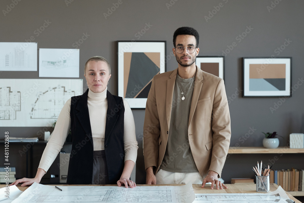 Poster two contemporary engineers standing by table with sketch and looking at you against grey wall with a
