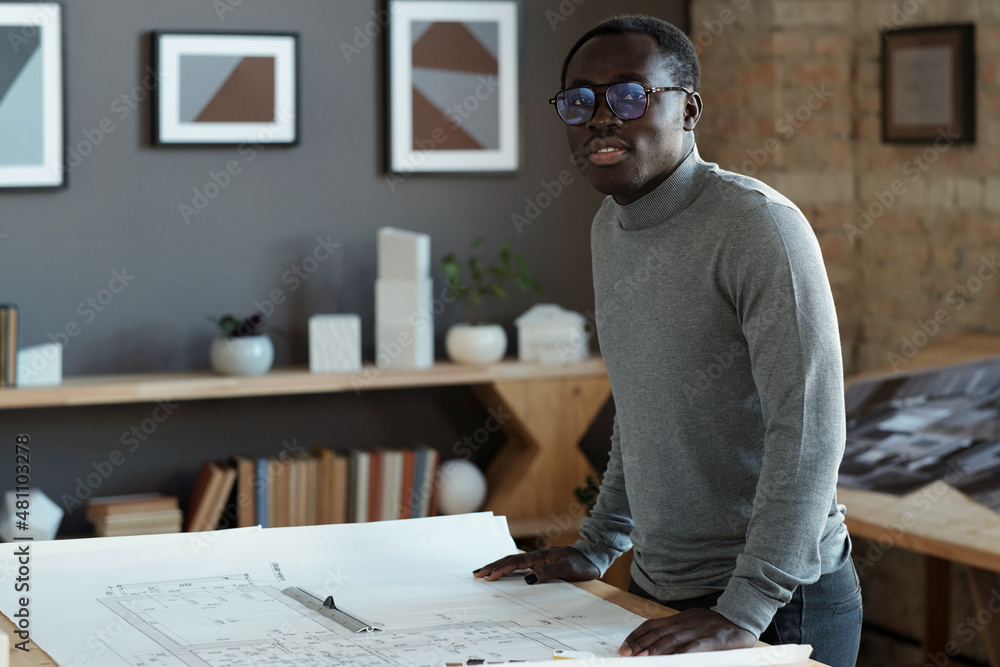 Aufkleber Contemporary young architect in eyeglasses and grey pullover looking at camera while standing by table with blueprint
