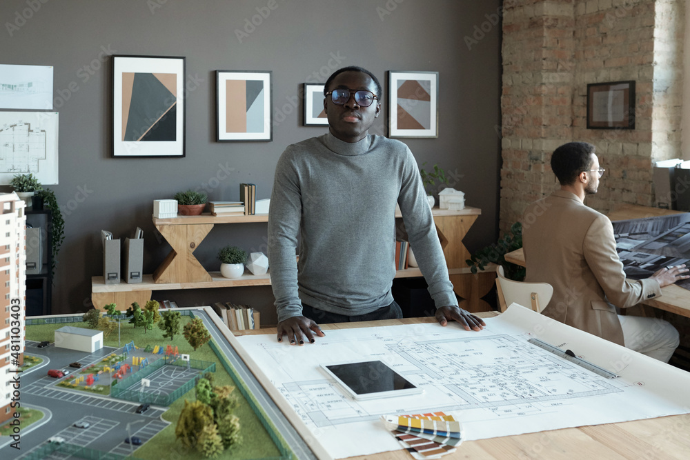 Poster Young African engineer standing by table with sketch and house layout and looking at you while working over new project