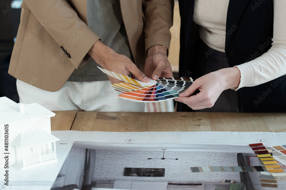 Poster Hands of two designers of interior choosing color for apartment walls while standing by table with blueprint and photos