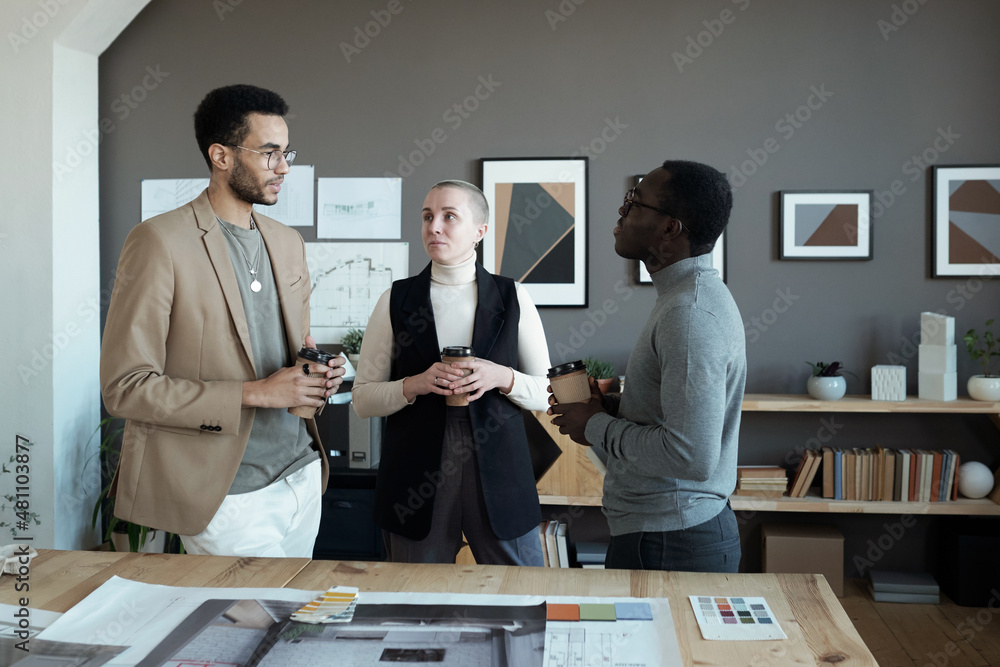 Poster Three creative designers discussing working points while standing by workplace and having coffee at break