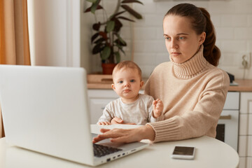 Indoor shot of serious concentrated woman with ponytail hairstyle wearing casual style beige sweater sitting at table in kitchen with infant kid, trying to work, needs finish work, deadline.