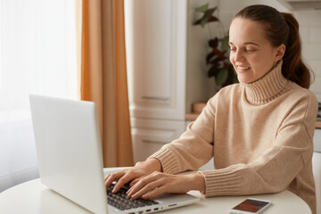 Portrait of positive attractive Caucasian woman wearing beige casual style sweater with ponytail hairstyle sitting at table and typing text on portable computer, expressing optimism.