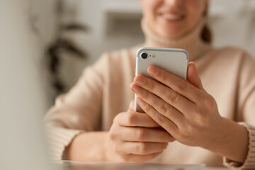 Closeup portrait of woman wearing beige casual style sweater using cell phone, watching videos in social networks or typing e-mail, expressing positive emotions, smiling.