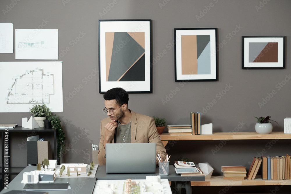 Poster Contemporary young pensive architect looking at layout of block of flats while sitting by table in front of laptop in office