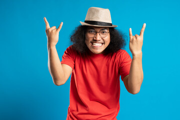 young man standing over blue background shouting with crazy expression doing rock symbol with hands up