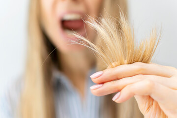 A shocked and upset blond woman holds the damaged brittle dry split ends of her long hair in her hand in front of her face, her mouth wide open in surprise, close-up. Health care and haircare concept. - obrazy, fototapety, plakaty