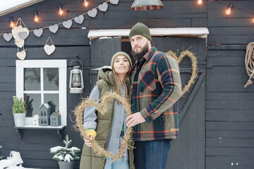 young couple in love beautiful Asian girl and bearded man in stylish winter clothes standing near porch of country house with handmade wreath of dried grass in shape heart, winter spirit valentine day