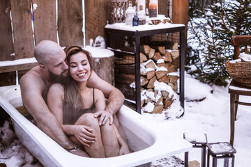 Obraz na płótnie Canvas young couple in love beautiful Asian girl and bearded man sit in bath with hot water on the snow near porch of country house with wooden fence decorated to celebrate Valentine's Day in suburbs