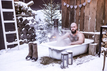 young brutal bearded tattoo man sit in bath with hot water on the snow near porch of country house...