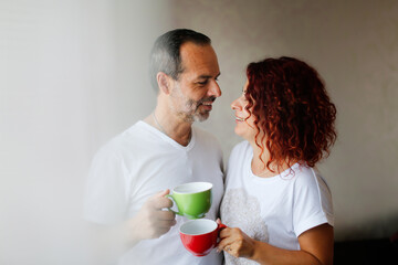 Cute middle aged european couple tender hugs and morning coffee together. Man and woman together at the window in the bedroom, lifestyle in a real interior. Support and emotions
