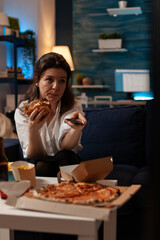 Woman sitting on sofa eating tasty delicious burger holding remote control switching channels on television in front of table with large pizza. Person browsing tv stations while eating hamburger.