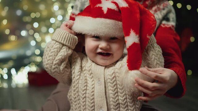 Mom puts a red Christmas hat on her baby. Family Spending Winter Evening at Home. Young Mother with Little Kid Son play. Slow Motion. Christmas, New Year, Winter Holiday Celebration. 4K, 10 bit colors