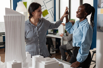 Team of two women architects high fiving standing in front of table with foam scale model of residential buildings in architectural modern office. Successful happy engineers celebrating team project.