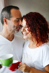 Close-up of a cute European middle-aged couple, tenderness and hugging. Care and emotions. Man and woman together at the window in the bedroom, lifestyle in a real interior.