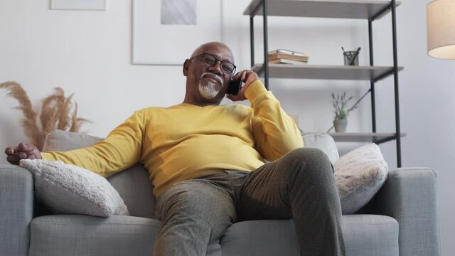 Pleasant communication. Happy senior man. Relaxed african aged guy talking by smartphone sitting comfortable sofa in light room interior.