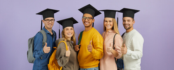 Education and success: Happy confident multiracial male and female university students, classmates and best friends in graduate hats give thumbs up standing together on purple color studio background