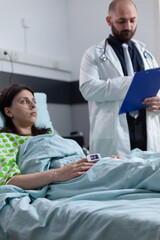Fototapeta premium Doctor with clipboard reading medical data to patient with nasal cannula recieving oxygen in hospital bed. Patient having lung disease with pulse oximeter on finger showing low spo2 saturation.
