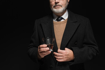 partial view of senior man in stylish coat holding glass of whiskey isolated on black.