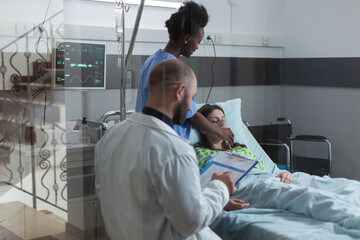 Fototapeta premium Through the window view of nurse putting oxygen mask on unconscious female patient in modern hospital ward. Consulting doctor writing on clipboard with medical lab results while caregiver attends.