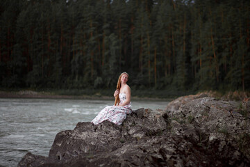 blonde with long hair, a girl walks by a mountain river before the rain, a gloomy heavy sky, clouds, the Katun River, the Altai Mountains, an art photo session, a dress with a floral print