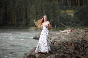 Fototapeta na wymiar blonde with long hair, a girl walks by a mountain river before the rain, a gloomy heavy sky, clouds, the Katun River, the Altai Mountains, an art photo session, a dress with a floral print