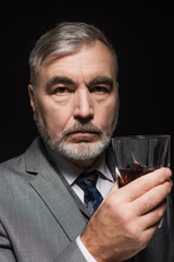 senior bearded man in blazer and tie holding glass of whiskey isolated on black.