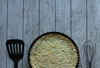 pie crumbly with cottage cheese in black form on a wooden background with a spatula and a whisk